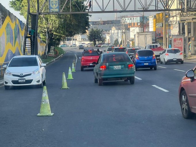 Street scene in an urban area of Villa Nueva with multiple vehicles including white Toyota sedans, red cars, green Honda, blue SUV, and pink vehicle driving on a two-lane road marked with white lines. Orange traffic cones are placed along the right side to guide flow near the reversible lane setup. Background shows buildings with colorful murals, trees, a metal bridge structure overhead, and a sign reading MOLINOS on a yellow building.