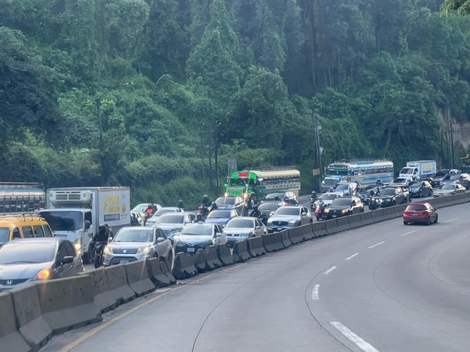 A multi-lane highway curves through lush green forested hills under daylight with heavy traffic congestion featuring numerous cars in various colors including white yellow and red trucks buses and motorcycles stopped or slowly moving along barriers and guardrails a green bus is visible among the vehicles and distant trees line the background.