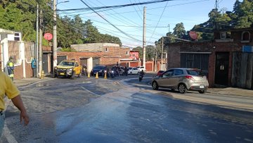 First image displays four workers in yellow vests and helmets walking along a wet roadside near a hillside with a no parking sign and vegetation power lines overhead and buildings in the background. Second image similar to first showing workers approaching a sloped earth bank with a no parking sign cars parked nearby and urban structures. Third image shows a street intersection with multiple vehicles including a yellow taxi and gray car traffic cones blocking access and a person in yellow vest near brick buildings and trees. Fourth image depicts excavators and machinery clearing large rocks and debris from a landslide area with a sign reading Escuintla and protective netting on a slope.