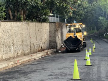 First image displays a yellow asphalt paving machine actively spreading material on a narrow street lined with green safety cones and a tall beige concrete wall covered in greenery including trees and vines in the background under overcast skies. Second image captures the same yellow machine from a side angle with its front blade lifted dumping asphalt onto the road surface surrounded by additional green cones and partial views of adjacent buildings and foliage. Third image shows several officers in high-visibility green vests and helmets standing on a wet asphalt road directing traffic with orange plastic barriers white vans and a yellow school bus visible nearby along with traffic cones and distant trees.