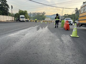 First image displays a yellow asphalt paving machine actively spreading material on a narrow street lined with green safety cones and a tall beige concrete wall covered in greenery including trees and vines in the background under overcast skies. Second image captures the same yellow machine from a side angle with its front blade lifted dumping asphalt onto the road surface surrounded by additional green cones and partial views of adjacent buildings and foliage. Third image shows several officers in high-visibility green vests and helmets standing on a wet asphalt road directing traffic with orange plastic barriers white vans and a yellow school bus visible nearby along with traffic cones and distant trees.