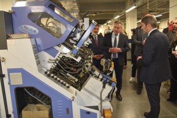 First image shows a yellow building facade with Olgunlasma Enstitusu Konya sign and logo, featuring a group of four people standing outside including women in dresses holding bouquets and men in suits. Second image depicts a pink building entrance with Konya Meram Halk Egitimi Merkezi sign and logo, showing a large group of about 15 people in professional attire posing on steps in front of the building with banners. Third image displays a blue industrial sewing machine in an indoor setting with several people in suits and dresses observing it closely. Fourth image portrays an indoor room with wooden ceiling and table covered in fabrics, yarns, crafts, and books, surrounded by a group of people including women in headscarves and men in suits engaged in activities.