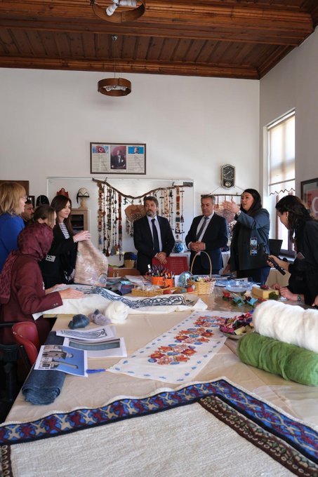 First image shows a yellow building facade with Olgunlasma Enstitusu Konya sign and logo, featuring a group of four people standing outside including women in dresses holding bouquets and men in suits. Second image depicts a pink building entrance with Konya Meram Halk Egitimi Merkezi sign and logo, showing a large group of about 15 people in professional attire posing on steps in front of the building with banners. Third image displays a blue industrial sewing machine in an indoor setting with several people in suits and dresses observing it closely. Fourth image portrays an indoor room with wooden ceiling and table covered in fabrics, yarns, crafts, and books, surrounded by a group of people including women in headscarves and men in suits engaged in activities.