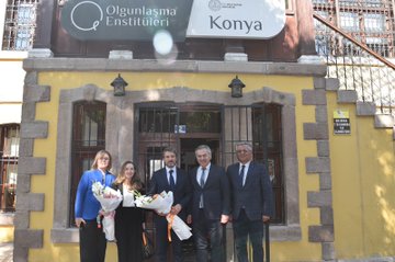 First image shows a yellow building facade with Olgunlasma Enstitusu Konya sign and logo, featuring a group of four people standing outside including women in dresses holding bouquets and men in suits. Second image depicts a pink building entrance with Konya Meram Halk Egitimi Merkezi sign and logo, showing a large group of about 15 people in professional attire posing on steps in front of the building with banners. Third image displays a blue industrial sewing machine in an indoor setting with several people in suits and dresses observing it closely. Fourth image portrays an indoor room with wooden ceiling and table covered in fabrics, yarns, crafts, and books, surrounded by a group of people including women in headscarves and men in suits engaged in activities.