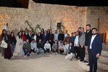 First image shows a large diverse group of about 30 young adults and professionals of various ages and genders dressed in casual to semi-formal attire including jackets dresses and shirts posing together outdoors at dusk against a lit ancient stone wall with decorative plants and the word MARDIN spelled in lights. Second image depicts another group of around 25 participants sitting and standing in a stone-walled room with arched doorways bookshelves colorful boxes and educational posters on Erasmus+ sitting on a red carpeted floor during an indoor event. Third image features four men in suits and traditional seating on red cushions around low tables with cups and plates in a stone room with bookshelves plants and banners discussing intently. Fourth image captures two suited men one gesturing with hand on chest walking up stone stairs in a warmly lit stone corridor with another person in background during the event.