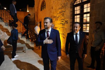 First image shows a large diverse group of about 30 young adults and professionals of various ages and genders dressed in casual to semi-formal attire including jackets dresses and shirts posing together outdoors at dusk against a lit ancient stone wall with decorative plants and the word MARDIN spelled in lights. Second image depicts another group of around 25 participants sitting and standing in a stone-walled room with arched doorways bookshelves colorful boxes and educational posters on Erasmus+ sitting on a red carpeted floor during an indoor event. Third image features four men in suits and traditional seating on red cushions around low tables with cups and plates in a stone room with bookshelves plants and banners discussing intently. Fourth image captures two suited men one gesturing with hand on chest walking up stone stairs in a warmly lit stone corridor with another person in background during the event.