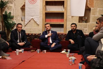 First image shows a large diverse group of about 30 young adults and professionals of various ages and genders dressed in casual to semi-formal attire including jackets dresses and shirts posing together outdoors at dusk against a lit ancient stone wall with decorative plants and the word MARDIN spelled in lights. Second image depicts another group of around 25 participants sitting and standing in a stone-walled room with arched doorways bookshelves colorful boxes and educational posters on Erasmus+ sitting on a red carpeted floor during an indoor event. Third image features four men in suits and traditional seating on red cushions around low tables with cups and plates in a stone room with bookshelves plants and banners discussing intently. Fourth image captures two suited men one gesturing with hand on chest walking up stone stairs in a warmly lit stone corridor with another person in background during the event.