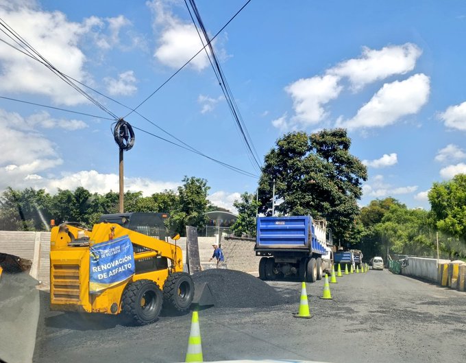 First image displays a yellow loader with a banner reading RenovaciÃ³n de Asfalto on its arm, positioned near a pile of asphalt on a road under construction, with a blue dump truck unloading material nearby, orange traffic cones lining the area, green trees and utility poles with wires overhead, and a clear blue sky with some clouds. Second image shows several workers in yellow safety vests and helmets walking around a freshly paved asphalt area surrounded by orange cones, with yellow excavators and a white truck in the background, a chain-link fence, and more trees under a blue sky. Third image captures a similar scene to the first, featuring the yellow loader, blue dump truck, asphalt pile, cones, trees, wires, and construction barriers on the roadside.