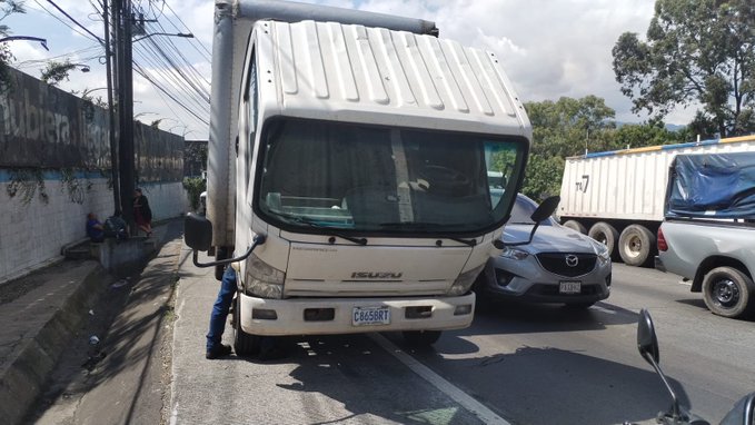 A white cab-over truck with Isuzu branding and license plate 489GAT is positioned on a sloped roadside, appearing immobilized with its front facing uphill. A person in dark pants stands near the drivers side door. Nearby, a gray Mazda sedan is parked close to the truck, and another white truck with a covered trailer is visible in the background. Utility poles and a graffiti-covered wall line the street, with trees and a partly cloudy sky overhead.