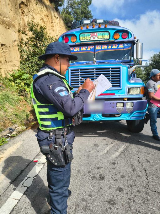 Un oficial de policia con uniforme azul y chaleco verde reflectante usa sombrero azul marino y escribe en una libreta junto a un autobus escolar colorido azul con detalles amarillos y texto en el frente que dice PREESCOLARIDE estacionado en una carretera empinada con bordes de tierra y vegetacion verde a los lados bajo un cielo nublado con otro oficial y una persona de pie cerca del vehiculo.