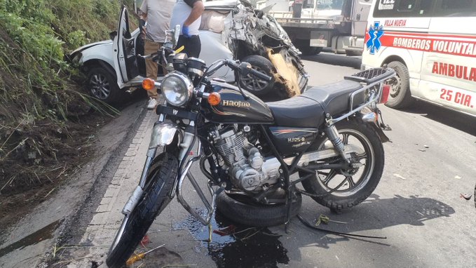 A black Honda motorcycle lies crashed on its side on a paved roadside with fluid leaking from it, next to a white car with its door open and a person standing nearby, a damaged truck in the background, and a white ambulance marked Bomberos Voluntarios 25 with emergency lights on parked adjacent to the scene amid greenery.