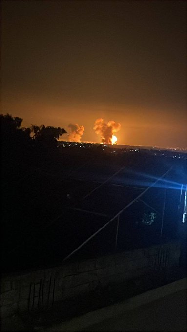First image displays a dark nighttime landscape with a bright orange glowing explosion resembling a sun on the horizon illuminating distant hills trees and scattered buildings under a hazy sky. Second image shows two large orange fiery blasts erupting from the ground amid a dark expansive terrain with silhouetted trees buildings and a lit-up structure in the foreground captured at night.