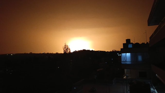 First image displays a dark nighttime landscape with a bright orange glowing explosion resembling a sun on the horizon illuminating distant hills trees and scattered buildings under a hazy sky. Second image shows two large orange fiery blasts erupting from the ground amid a dark expansive terrain with silhouetted trees buildings and a lit-up structure in the foreground captured at night.