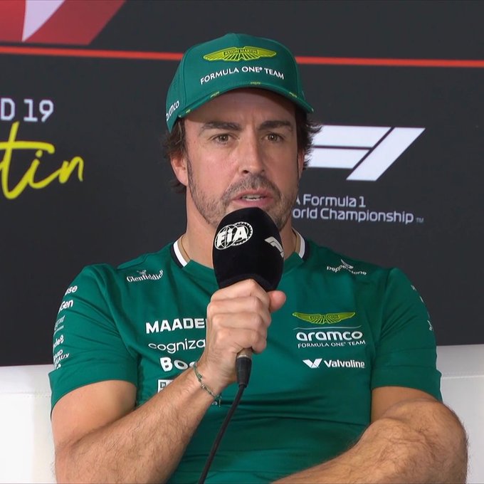 Fernando Alonso sits in a press conference wearing a green Aston Martin Formula One Team cap and matching team shirt with sponsor logos including Aramco and Valvoline. He holds a black microphone labeled FIA World Championship and speaks into it. The background features a black banner with white text for Formula 1 and USGP 2019 branding.