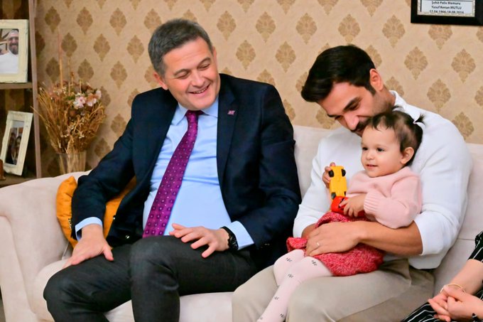 First image displays three adults sitting on a beige sofa in a warmly decorated living room with ornate wallpaper, framed photos, Turkish flags, and award plaques on walls; the man in the center wears a white shirt and light pants, the woman to his right a black patterned dress, and the man to the left a blue suit with purple tie. Second image shows a man in a dark blue suit with purple tie smiling and interacting with a smiling infant girl in pink clothing held by a young man in white shirt next to him, seated on a sofa with beige cushions and flower vase nearby in a similar room setting.