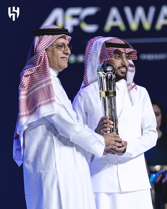 Two men in traditional Saudi attire one older with glasses wearing a red-and-white shemagh over white thobe and younger man with similar attire both smiling and standing close together on a stage holding a large shiny silver trophy with blue base and gold accents the older man grips the base while younger holds the top background features dark blue backdrop with white AFC Champions League logo and event branding no other objects visible