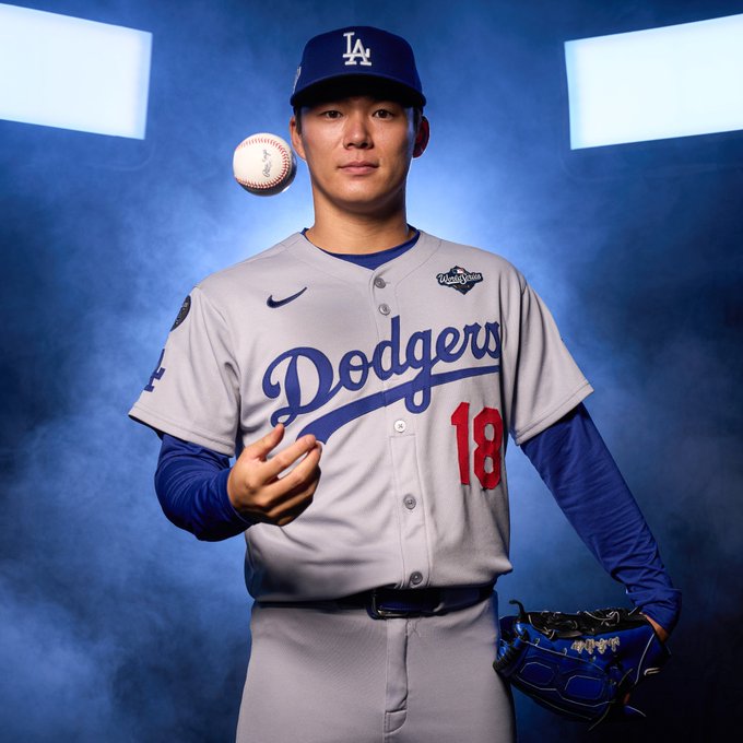 Yoshinobu Yamamoto tosses a baseball and poses during the World Series photoshoot wearing a gray Los Angeles Dodgers uniform with blue lettering and red numbering.