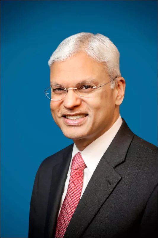 Portrait of an elderly Indian man with white hair, wearing thin-rimmed glasses, smiling slightly, dressed in a gray suit with a red striped tie, posed against a solid blue background.