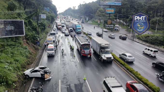 Aerial view of a multi-lane highway with heavy traffic including multiple large trucks cars motorcycles and buses moving slowly in rainy conditions surrounded by green hills and urban buildings with a visible PMT Villa Nueva police badge emblem on a signpost and several police officers in black uniforms and helmets standing along the roadside directing traffic near the congestion area.