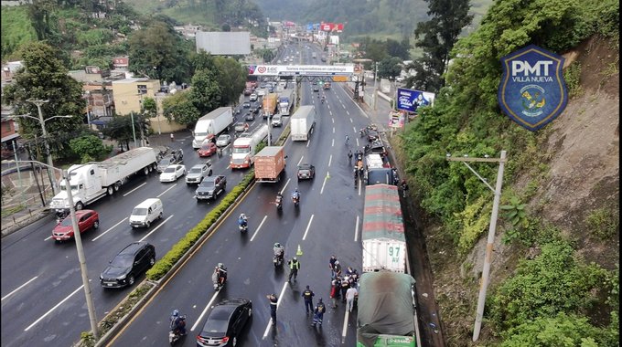 Aerial view of a multi-lane highway with heavy traffic including multiple large trucks cars motorcycles and buses moving slowly in rainy conditions surrounded by green hills and urban buildings with a visible PMT Villa Nueva police badge emblem on a signpost and several police officers in black uniforms and helmets standing along the roadside directing traffic near the congestion area.