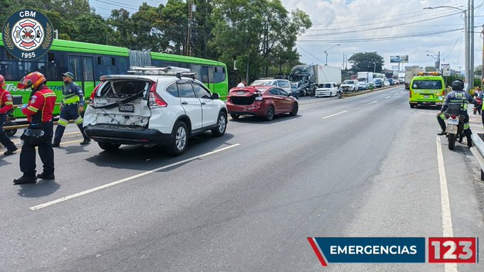 Scene on a multi-lane road with trees and overhead signs showing a green bus on the left side damaged white SUV with rear-end collision red sedan with front damage white van and other vehicles stopped emergency responders in red uniforms and helmets attending with a yellow motorcycle nearby green text overlay EMERGENCIAS and number 123