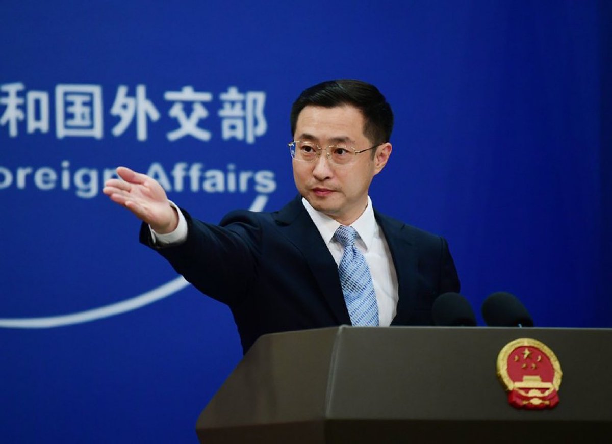 Man in dark suit and blue tie stands at wooden podium with microphones gesturing with right hand during speech blue backdrop displays Foreign Affairs in Chinese characters national emblem on podium side