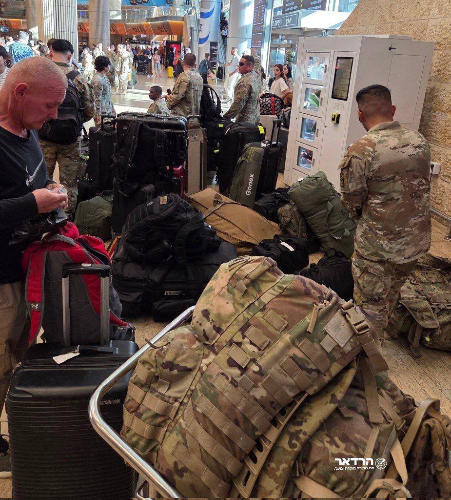 Crowded airport terminal with multiple people including men in military uniforms carrying backpacks and bags near luggage carts and a vending machine. Hebrew text visible on signs and displays. Soldiers in camouflage attire stand among civilian passengers with suitcases and duffel bags on the floor.