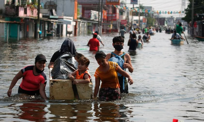 People wade through knee-deep muddy floodwaters on a street lined with colorful buildings and hanging banners. Several individuals carry a large wooden box with a child inside, while others push small boats or walk with belongings. The scene shows urban flooding with vehicles partially submerged and residents navigating the water.