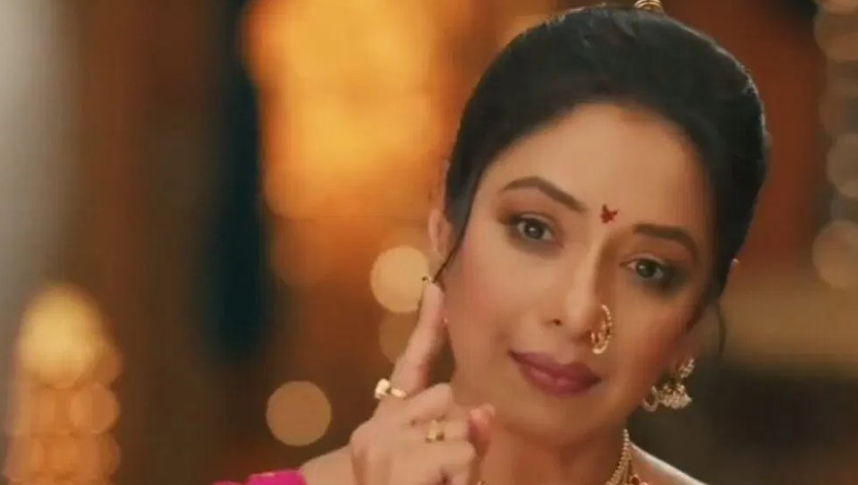 A woman with black hair styled in a bun and wearing a red bindi on her forehead, a gold nose ring, and large gold earrings stands in a warmly lit setting with blurred orange background lights. She has kohl-lined eyes, red lips, and a subtle smile while raising her right hand with fingers extended near her face. She wears a pinkish-red outfit with gold necklace and visible gold bracelet on her wrist.
