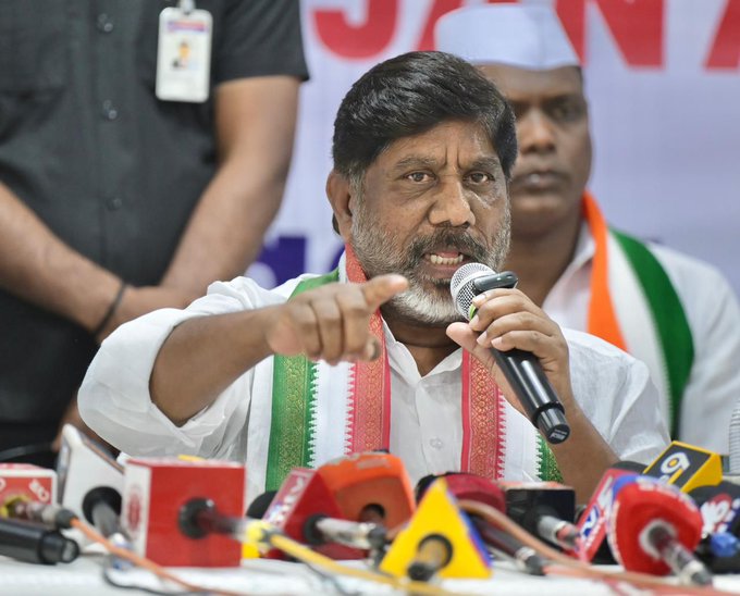 A man in a white shirt with green-bordered saffron scarf around his neck holds a microphone and points forward while speaking at a press conference. He has a mustache and wears a white cap. Behind him stand two men in dark shirts one with a badge and another in orange attire. A banner with orange and white colors is visible in the background. Multiple microphones from various media outlets including red and yellow ones are placed on a table in front of him.