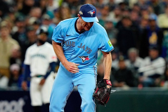 A male baseball player wearing a blue Toronto Blue Jays cap and uniform jersey with the team logo stands on a baseball field during a game. He has a red glove on his right hand and is positioned near the outfield with a stadium crowd and other players in the background. The uniform includes blue pants and a green patch on the sleeve.