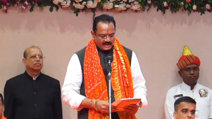 A man in orange scarf and white attire stands at a podium holding a book and microphone, speaking during a ceremony. He wears glasses and has garlands around his neck. Other men in formal clothes, one in black suit and another in uniform with red turban, stand beside him. Background features pink wall with flower garlands.
