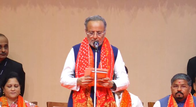 An elderly man with glasses and gray hair stands at a podium holding a book wearing a white kurta with blue and red scarf over it speaking into a microphone. He is flanked by other men and women in formal Indian attire including kurtas and sarees some with red and blue scarves seated on a stage with orange backdrop and garlands.