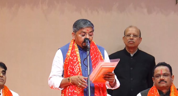 A man with gray hair wearing a white kurta, orange scarf, and vest stands at a podium holding a document and microphone. He is flanked by three other men in formal attire including white kurtas, black suits, and orange scarves. The setting is a stage with a beige backdrop and microphone stand.