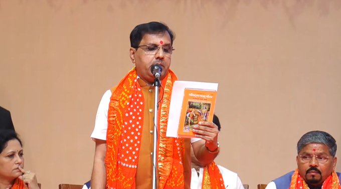 A man in orange traditional attire and white shirt stands at a podium holding a microphone and an orange book with text on it. He has a red mark on his forehead and wears a wristband. Two other men in similar orange attire sit nearby one with glasses and a mustache. A woman in a maroon outfit sits beside them. The background is a plain wall with a small emblem.