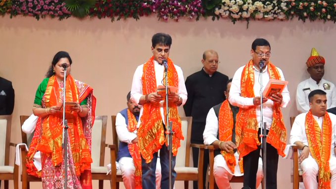 Group of politicians on a stage with floral decorations in background. Central figures include a woman in green saree holding a plate, a man in white kurta with saffron scarf speaking into microphone, another man in orange attire holding a book, and others in formal suits and turbans seated or standing nearby. All appear engaged in a formal ceremony.