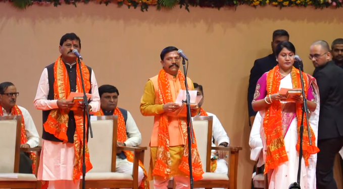 Three men and one woman stand on a stage during an oath-taking ceremony. The first man wears a white kurta with saffron shawl and holds a book. The second man in saffron kurta speaks into a microphone holding a book. The woman in pink saree with bangles holds a book. The third man in saffron attire stands nearby. Background includes beige wall, garlands, chairs, and other suited individuals.
