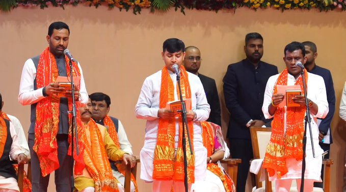 Group of men in traditional Indian attire including saffron scarves and white kurtas stand at podiums holding books, with some wearing formal suits, against a backdrop of orange walls and floral decorations in a ceremonial hall setting.