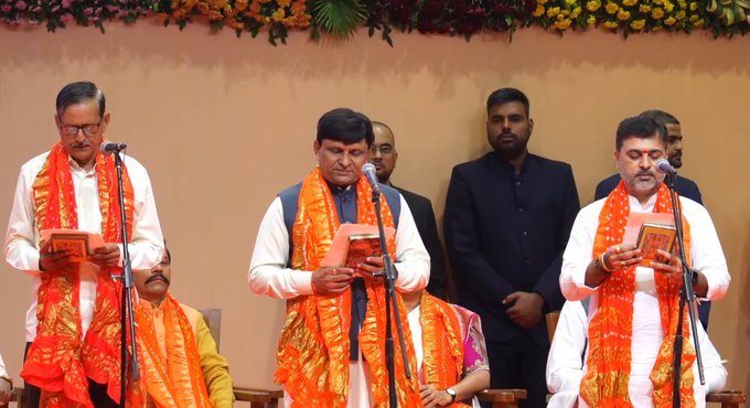 Three men in white kurtas and saffron scarves stand on a stage holding books and microphones during an oath ceremony. The stage features orange marigold garlands and drapes against a beige wall. Additional men in formal attire including suits and kurtas are visible nearby. The setting appears formal with decorative elements.