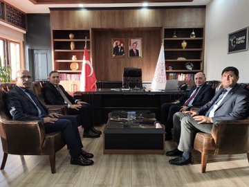 First image shows four men in suits seated around a wooden desk in an office with bookshelves, portraits on walls, Turkish and white flags, and decorative vases; they appear to be in a formal discussion. Second image depicts a long conference table with approximately 20 people including men and women in business attire seated around it, with Turkish flags, portraits, and projection equipment in a modern meeting room. Third image features a group of men in suits at a conference table with laptops, plates, and a projector screen displaying charts in a brightly lit room with clocks and windows. Fourth image illustrates several individuals including men and women in formal clothing around a wooden table with laptops and a projector screen showing a red logo in a room with stone walls, arched doorways, and traditional decor.