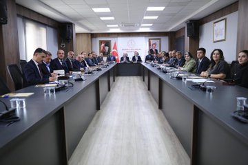 First image shows four men in suits seated around a wooden desk in an office with bookshelves, portraits on walls, Turkish and white flags, and decorative vases; they appear to be in a formal discussion. Second image depicts a long conference table with approximately 20 people including men and women in business attire seated around it, with Turkish flags, portraits, and projection equipment in a modern meeting room. Third image features a group of men in suits at a conference table with laptops, plates, and a projector screen displaying charts in a brightly lit room with clocks and windows. Fourth image illustrates several individuals including men and women in formal clothing around a wooden table with laptops and a projector screen showing a red logo in a room with stone walls, arched doorways, and traditional decor.