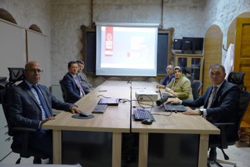 First image shows four men in suits seated around a wooden desk in an office with bookshelves, portraits on walls, Turkish and white flags, and decorative vases; they appear to be in a formal discussion. Second image depicts a long conference table with approximately 20 people including men and women in business attire seated around it, with Turkish flags, portraits, and projection equipment in a modern meeting room. Third image features a group of men in suits at a conference table with laptops, plates, and a projector screen displaying charts in a brightly lit room with clocks and windows. Fourth image illustrates several individuals including men and women in formal clothing around a wooden table with laptops and a projector screen showing a red logo in a room with stone walls, arched doorways, and traditional decor.