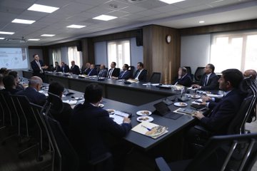 First image shows four men in suits seated around a wooden desk in an office with bookshelves, portraits on walls, Turkish and white flags, and decorative vases; they appear to be in a formal discussion. Second image depicts a long conference table with approximately 20 people including men and women in business attire seated around it, with Turkish flags, portraits, and projection equipment in a modern meeting room. Third image features a group of men in suits at a conference table with laptops, plates, and a projector screen displaying charts in a brightly lit room with clocks and windows. Fourth image illustrates several individuals including men and women in formal clothing around a wooden table with laptops and a projector screen showing a red logo in a room with stone walls, arched doorways, and traditional decor.