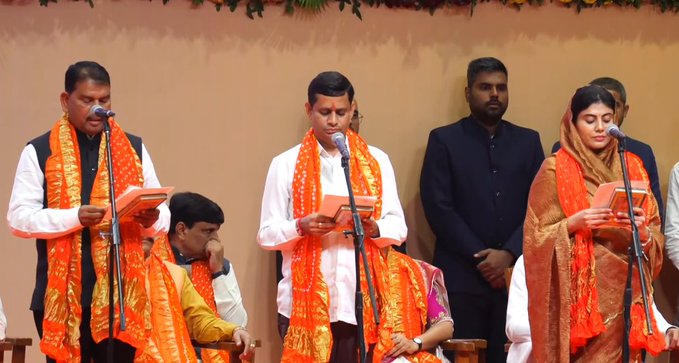 Group of people on a stage with beige background and green garlands, several men and women in orange and white traditional attire holding books and microphones, standing and seated positions during a formal ceremony.