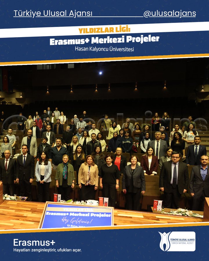 Group of people in formal business attire including suits jackets and dresses seated and standing in rows in a large auditorium with wooden paneling and stage. Front table displays banners for Erasmus+ Central Project and Hasan Kalyoncu University. University logo and event title Yıldızlar Ligi Erasmus+ Central Projects visible on backdrop. Attendees appear professional with some smiling at camera. Setting is indoors at Hasan Kalyoncu University event space.