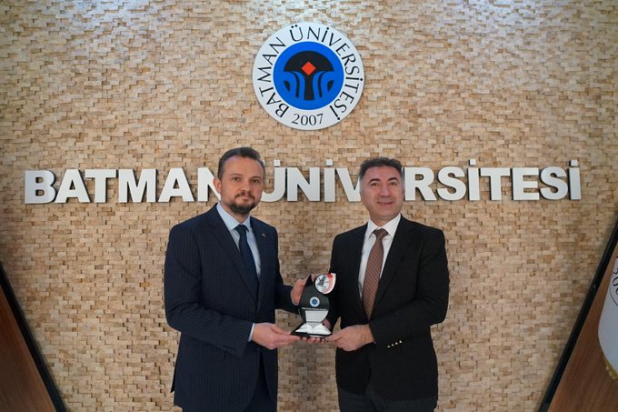 Two men in dark suits and ties stand in front of a beige tiled wall displaying the Batman University logo and Turkish flag on the left and an EU flag on the right in the first image. The wall has metallic lettering spelling Batman University. In the second image the same men hold a trophy between them with the university logo visible behind.