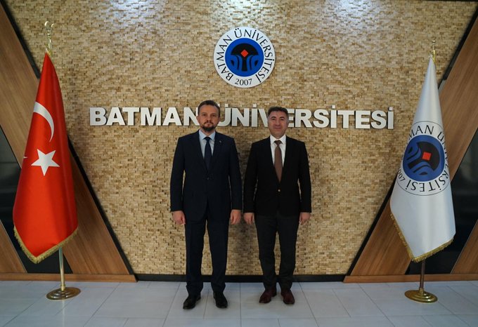 Two men in dark suits and ties stand in front of a beige tiled wall displaying the Batman University logo and Turkish flag on the left and an EU flag on the right in the first image. The wall has metallic lettering spelling Batman University. In the second image the same men hold a trophy between them with the university logo visible behind.