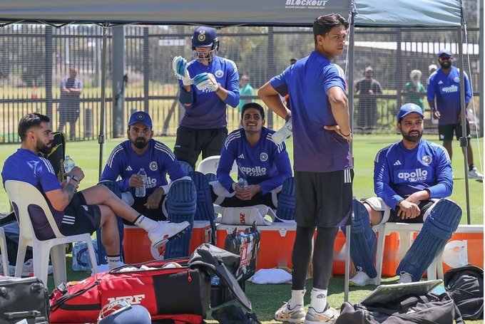 Group of male cricket players in blue uniforms including some with helmets gloves and bats gathered on a grassy field near a practice net enclosure white chairs scattered around equipment bags visible in the background sunny outdoor setting with a shaded pavilion structure