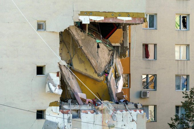 A multi-story apartment building in Bucharest shows extensive damage from an explosion, with the upper facade partially collapsed exposing interior rooms, twisted metal beams, hanging debris, and broken windows amid surrounding intact structures and some laundry on lines.