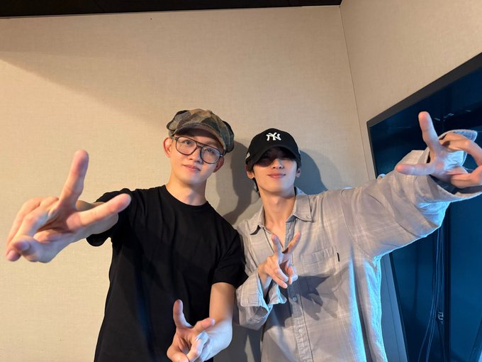Two young men standing side by side in a plain indoor room with beige walls and a dark door. The one on the left wears a black t-shirt and camo bucket hat with glasses making peace signs with both hands. The one on the right wears an oversized light gray shirt and black New York Yankees cap making peace signs with both hands smiling at the camera.
